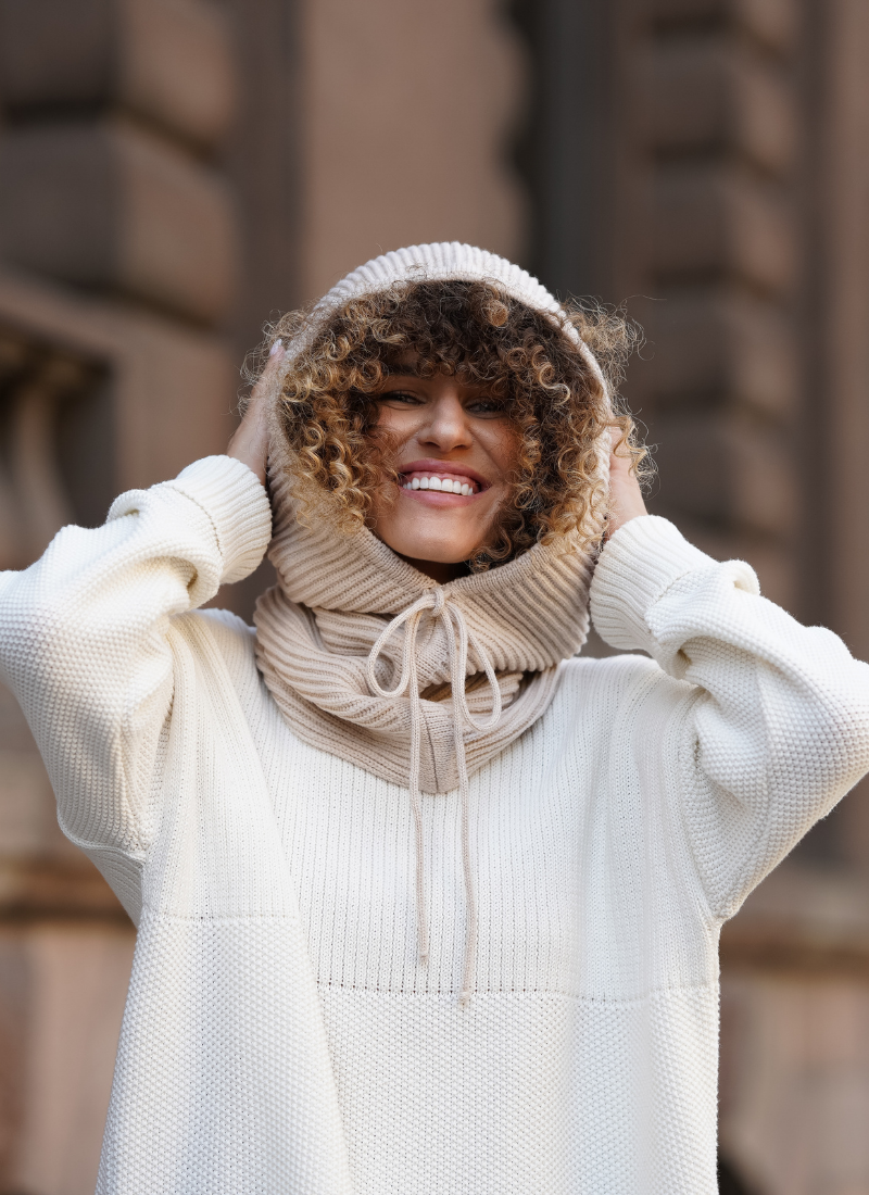 Smiling woman wearing a cream knit sweater and matching knit hooded scarf, standing outdoors in an urban setting, showcasing soft texture and cozy cold-weather style.