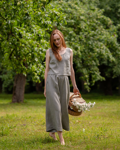 The model is visible from the front, looking down at the ground. She is wearing the LANA culottes in green gingham, while holding a basket full of wildflowers in her hand. Her bottoms are matched with a sleeveless blouse in the same pattern.