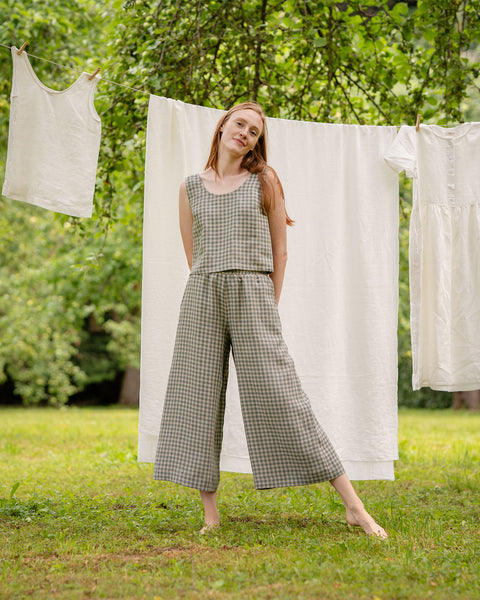 In this picture the model is posing full-height, showing the front of the green gingham culottes matched with the green gingham crop top. Behind her is a clothesline with white garments hanging on it. She is in a garden, under a tree.