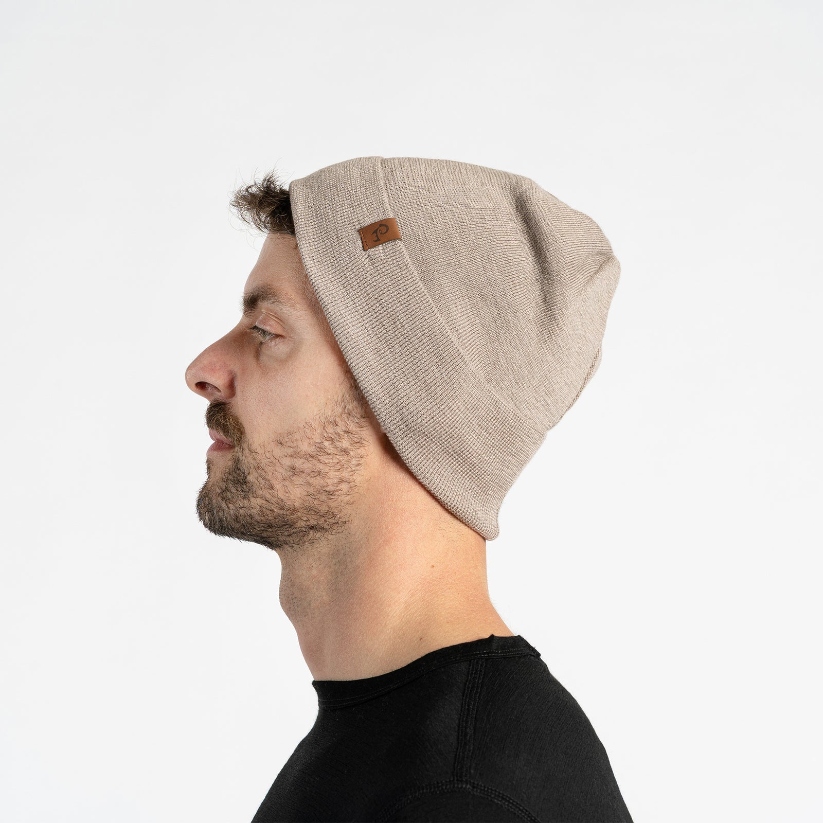 A man with light skin, blue eyes, and a beard smiles faintly while looking forward. He wears a creamy beige colored menique knit beanie with a small brown patch on the side, and a black crew-neck shirt.