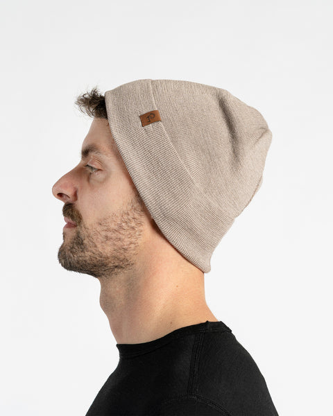 A man with light skin, blue eyes, and a beard. He wears a creamy beige colored menique knit beanie with a small brown patch on the side, and a black crew-neck shirt.