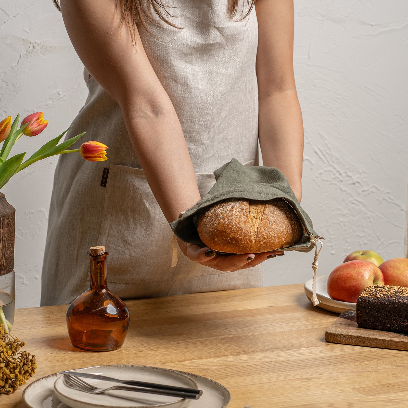 A studio laydown of two medium rectangular linen bread bags against a plain light gray background. One bag is olive green and the other is a natural beige color. Both feature natural rope drawstrings and small brown brand tags.