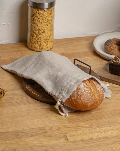 Loaf of bread wrapped in a cloth on a wooden table with a jar of pasta and bagels in the background.