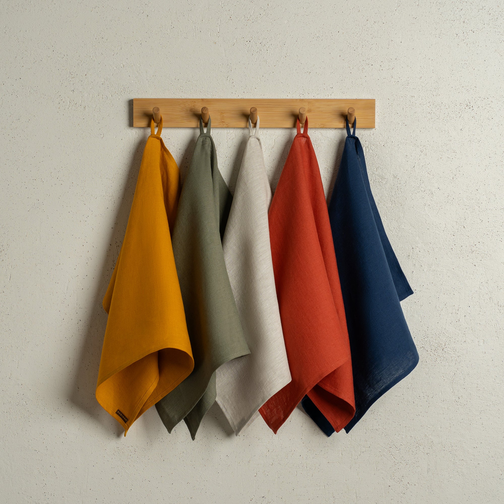 The menique linen kitchen dish towels in the colors of spicy yellow, stone green, natural, cinnamon red, and storm blue hanging on a wooden rack on a wall.