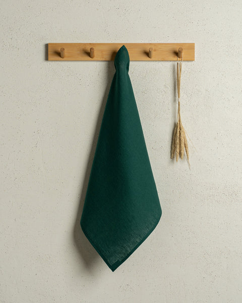 The menique square linen dish towel in color dark green hanging on a wooden rack on a wall.