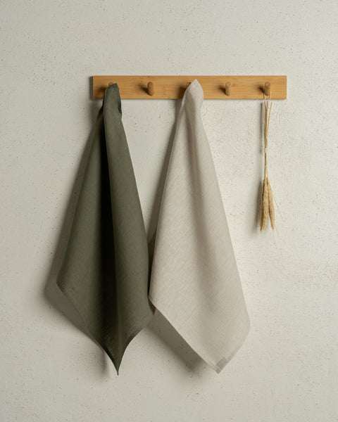 Two menique square linen dish towels in colors stone green and natural hanging on a wooden rack on a wall.