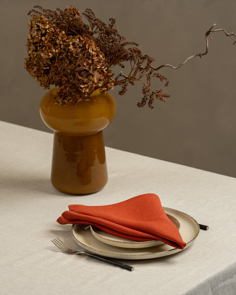 A tabletop covered with a linen tablecloth, with a vase and a single plate set on top of it. By the sides of the plates there are cutlery items, and there is a linen napkin in cinnamon red color on top.