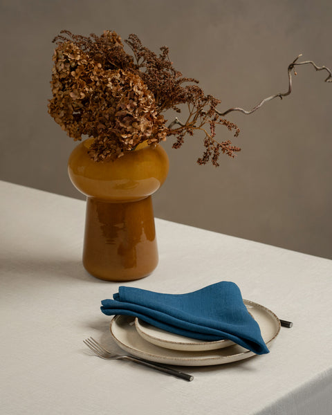 A tabletop covered with a linen tablecloth, with a vase and a single plate set on top of it. By the sides of the plates there are cutlery items, and there is a linen napkin in cobalt blue color on top.
