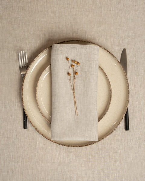The menique linen napkin in natural color, put on top of two stacked plates . There are cutlery items by the sides of the plates. The background is a natural color linen tablecloth.