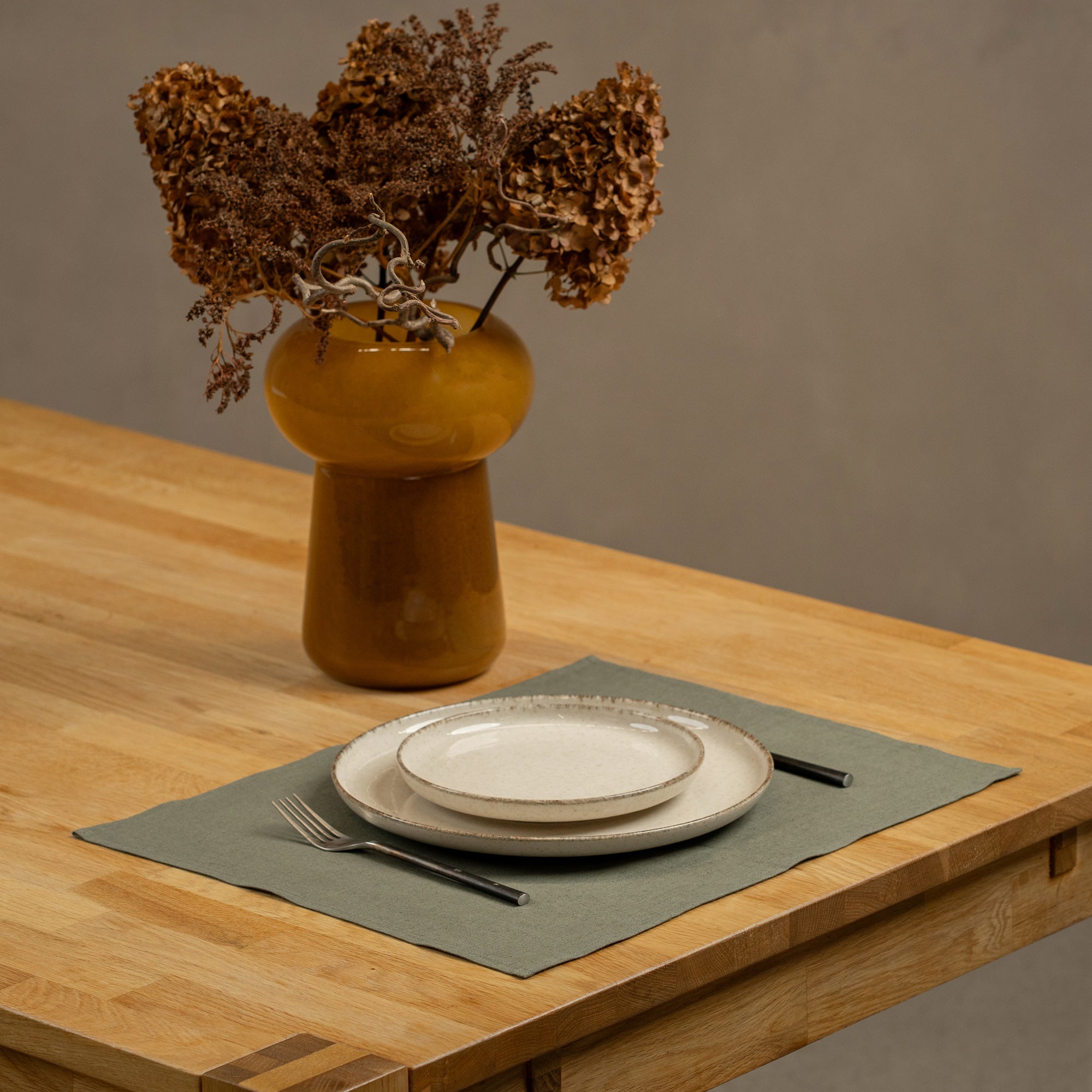 The menique cinnamon red linen placemat on a light wooden table. There are two different size porcelain plates and cutlery on top of it. There is also a vase on the table.