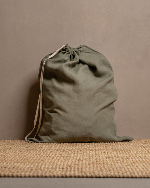 A closed, large stone green menique linen storage bag sits on a textured jute rug against a warm brown wall.