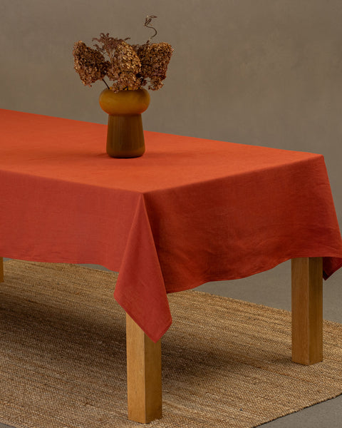 The menique cinnamon red linen tablecloth on a wooden table. On top of the tablecloth is a vase made of natural straws and a clay cup. Under the table is a carpet.