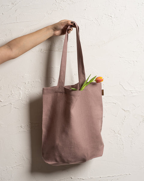 A fair-skinned arm and hand hold up a faded rose reusable linen tote bag against a textured off-white wall. A single tulip with a red and yellow bloom and green leaves is visible inside the bag.
