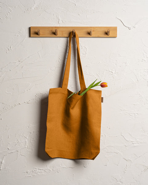 Menique spicy yellow durable linen tote bag hangs from a wooden rack with four pegs mounted on a textured off-white wall. 