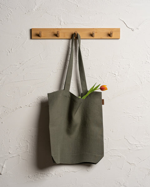 Menique reusable linen tote bag hangs from a wooden rack with four pegs mounted on a textured off-white wall. A single tulip with a red and yellow bloom and green leaves is visible sticking out of the top of the bag.