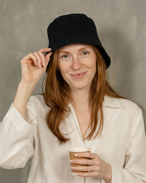 A smiling woman with red hair and freckles holding a paper coffee cup. She is gently touching the brim of her black bucket hat with her other hand. She is wearing a slightly wrinkled, white linen button-down shirt. The background is a textured, light grey wall.