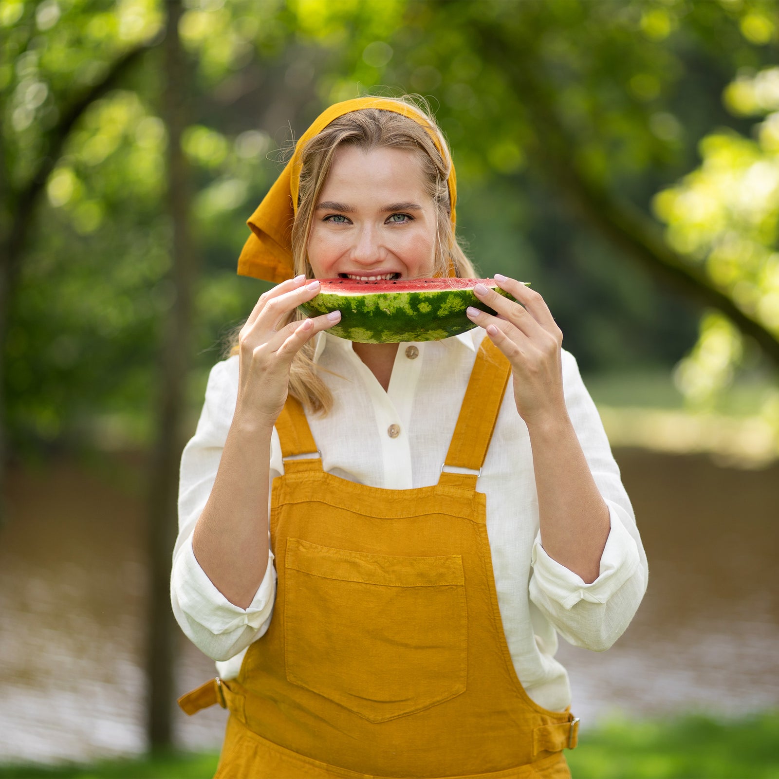 A blonde woman beams at the camera while standing outdoors against a backdrop of lush green trees. She rests both hands gracefully on top of her head while wearing a white shirt and brown skirt.
