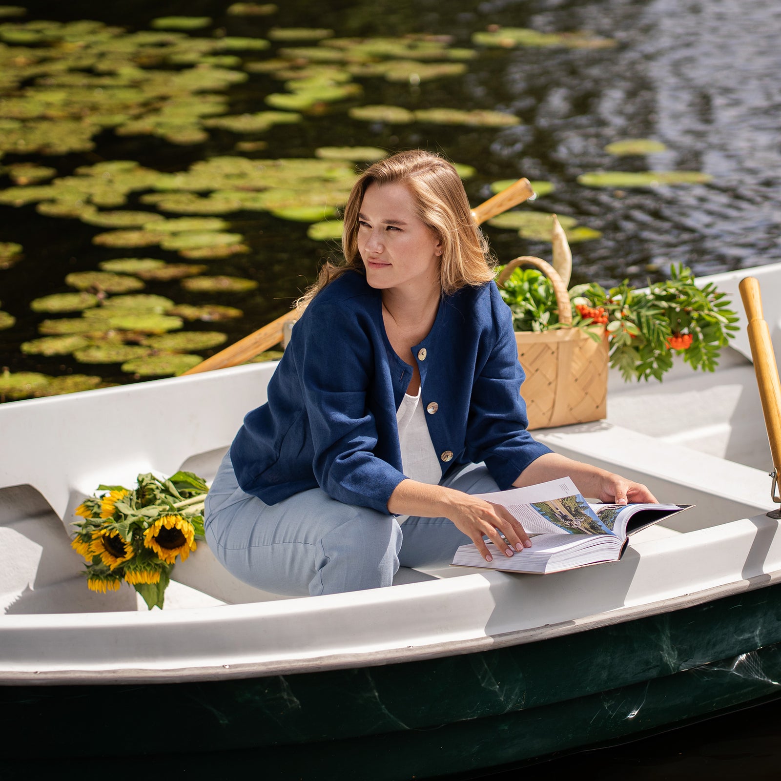 A woman wearing a blue jacket actively rows a small boat across a calm river. She holds the wooden oars firmly while looking forward and smiling.