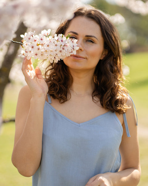 A woman wearing a cloudy blue linen slip dress stands beside a tree covered in white blossoms and gently smells a cluster of flowers.