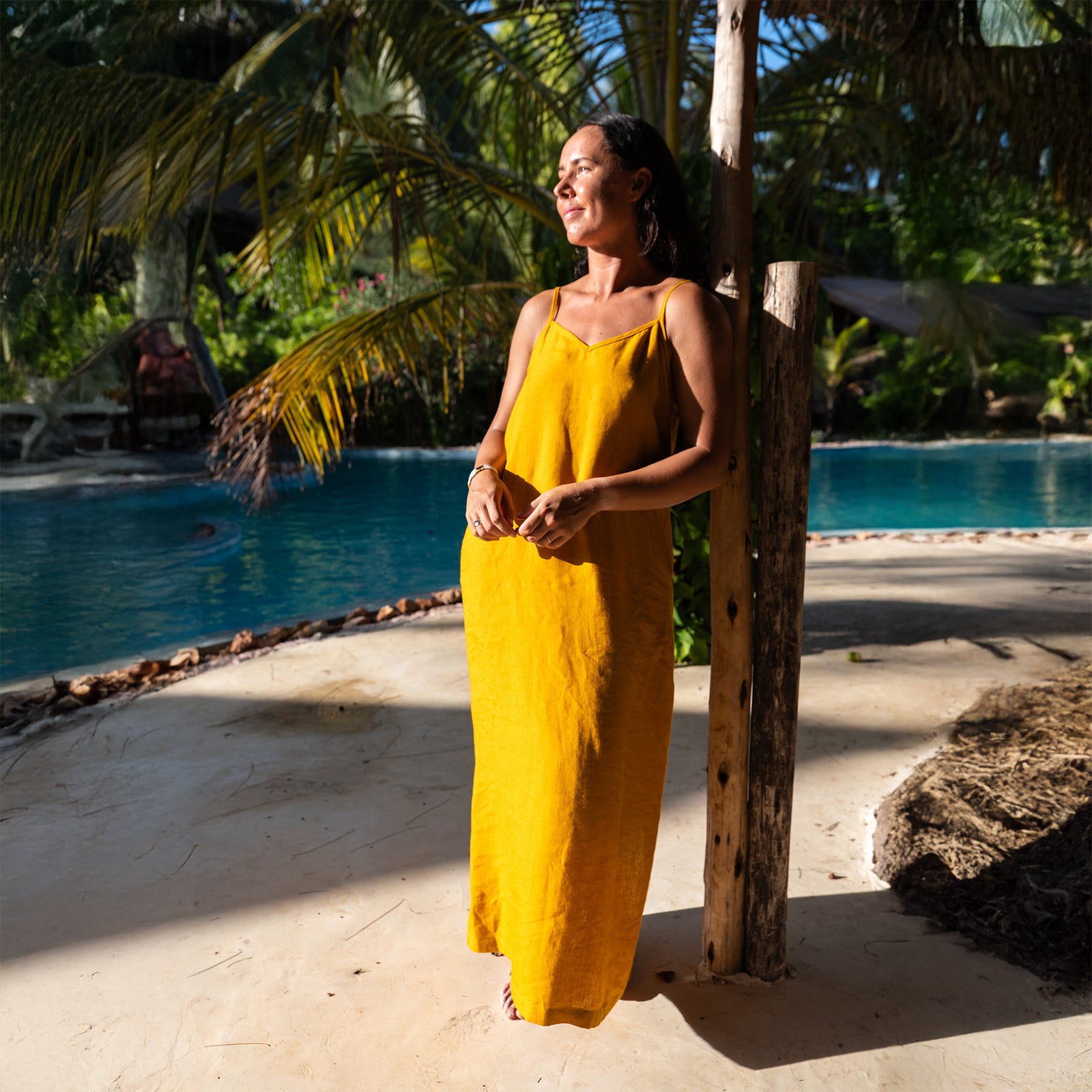 A woman with long, dark, wavy hair stands barefoot on a sandy path, looking up towards the left. She is wearing a mustard yellow linen sundress with thin straps. Lush green plants and trees surround her, and there are pink flowers blooming in the background. Yellow petals are scattered on the path.