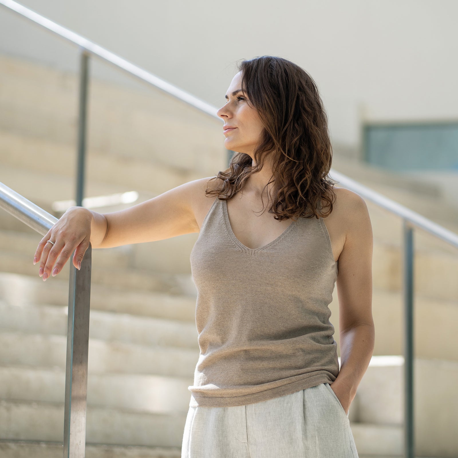 A woman with shoulder-length wavy brown hair stands looking upwards and to the right, with one arm resting on a silver handrail and the other hand near her forehead, as if shielding her eyes. She is wearing a creamy beige merino and cashmere tank top and light-colored linen trousers, and is positioned on or near a set of light-colored stairs with a glass or clear barrier. The lighting appears bright, possibly natural daylight.