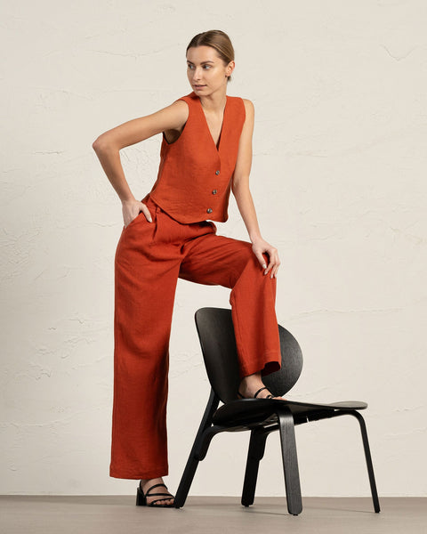 A woman in a cinnamon red colored linen vest and wide-leg pants poses with one leg resting on a black chair against a textured beige wall. She wears black sandals.
