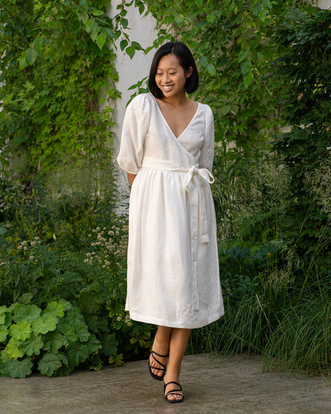 A smiling person with short, dark hair and what appears to be Asian features is standing on a paved surface surrounded by lush greenery. They are wearing a pure white wrap dress with elbow-length sleeves and a tied waist, along with black strappy sandals.