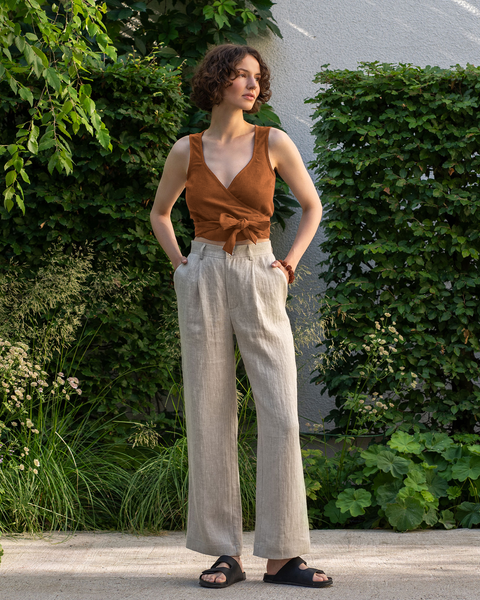 A light-skinned person with short, curly brown hair is standing outdoors with their hands in the pockets of light beige wide-legged pants. They are wearing an almond brown sleeveless wrap top tied at the side and dark slip-on sandals, and are looking to the right. Lush green foliage and a light-colored wall form the backdrop.