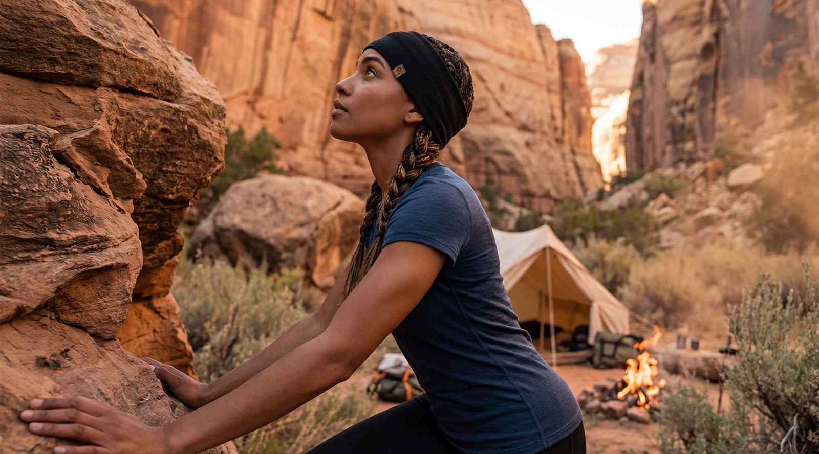 A side profile of a woman looking upwards as she leans against a large, textured red rock face. She has her hair in braids and is wearing a dark blue short-sleeved t-shirt and a wide black headband. A canvas tent is partially visible in the blurred background against the canyon walls.