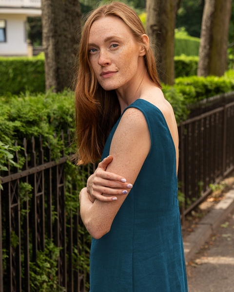 A woman with long reddish hair and freckles, wearing a menique linen sleeveless dress, stands with crossed arms on a sidewalk next to a black fence and green bushes.
