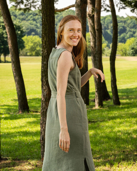 A fair-skinned woman with long, reddish-brown hair is captured from the waist up, smiling and looking back over her left shoulder towards the viewer. She is wearing a sleeveless, stone green linen dress with a tie belt around her waist. 