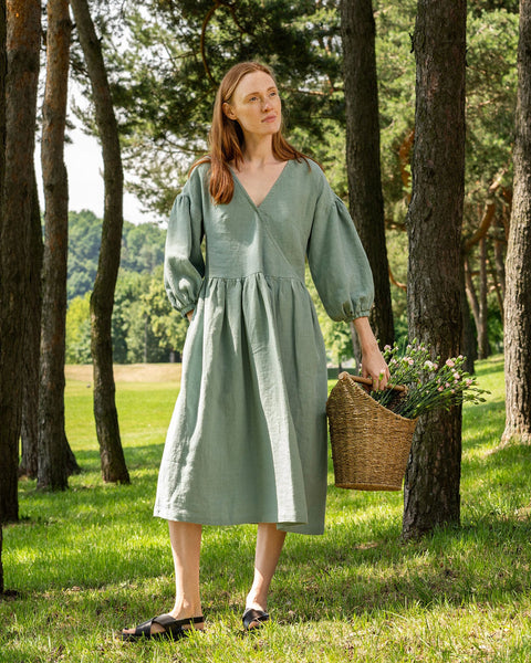 A full-body shot of a woman with long, reddish-brown hair standing in a grassy forest. She is looking upwards and to her right. She is wearing a knee-length, mint green linen wrap dress with a V-neck and elbow-length, puffed sleeves. She is holding a woven basket filled with white flowers and green foliage in her left hand. She is wearing black slides. 