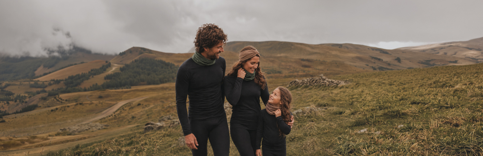 A family of three wearing matching black activewear stands together in a grassy mountain landscape under an overcast sky.