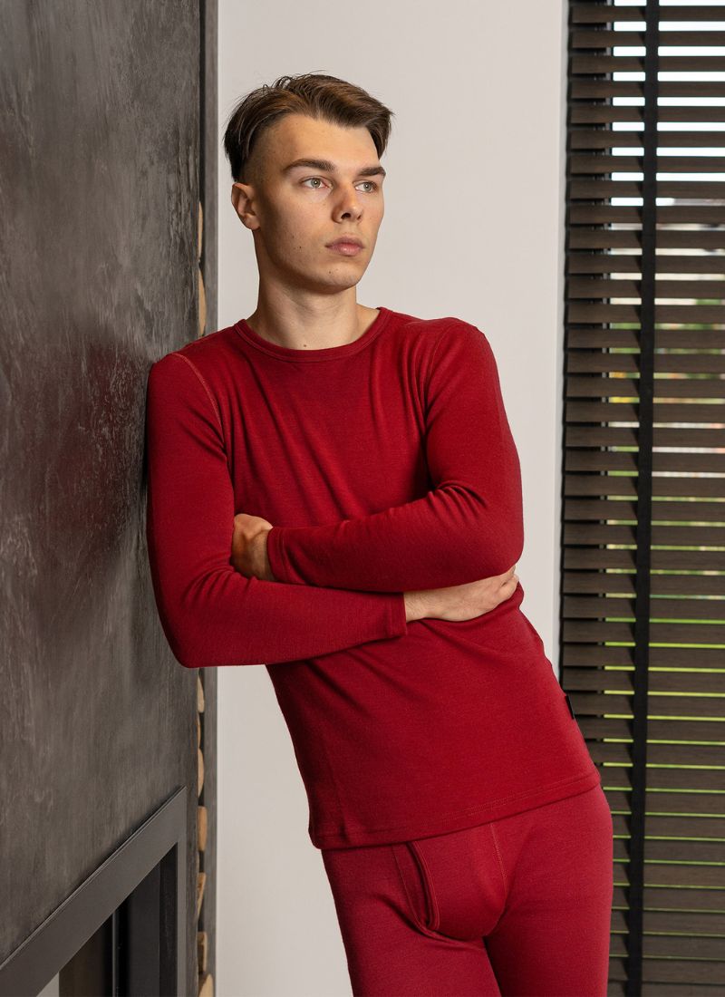 A young man leaning on a wall, wearing a merino wool long sleeve shirt and pants in royal cherry color.