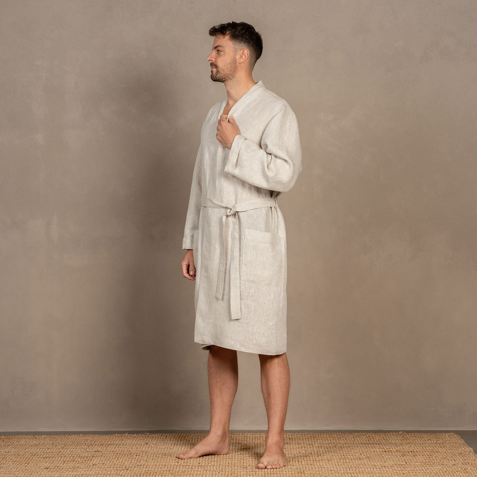 A man, turned slightly to the side, wearing the men's linen bathrobe in natural color. Both of his hands are in the side pockets.