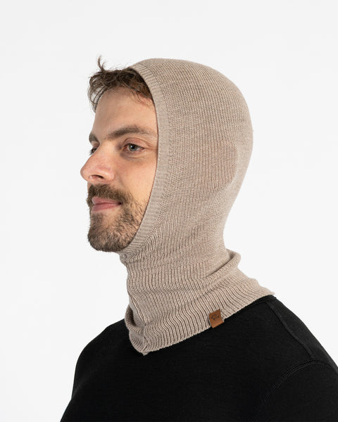 A man with a beard and short dark hair is shown in a profile shot, looking to his left. He is wearing a menique knit balaclava or hood that covers his head, ears, and neck, with a small brown leather tag visible on the lower left side. He is also wearing a black long-sleeved shirt. The background is plain white.