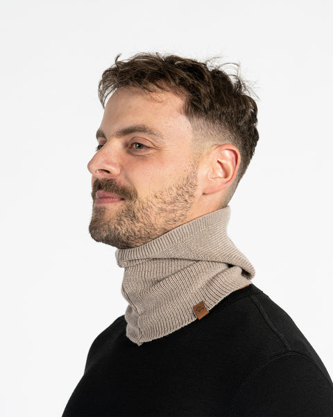 A man with short brown hair, a beard, and a mustache is seen in profile, wearing a creamy beige, ribbed  menique balaclava over a black long-sleeved shirt, looking off to the left with a slight smile.