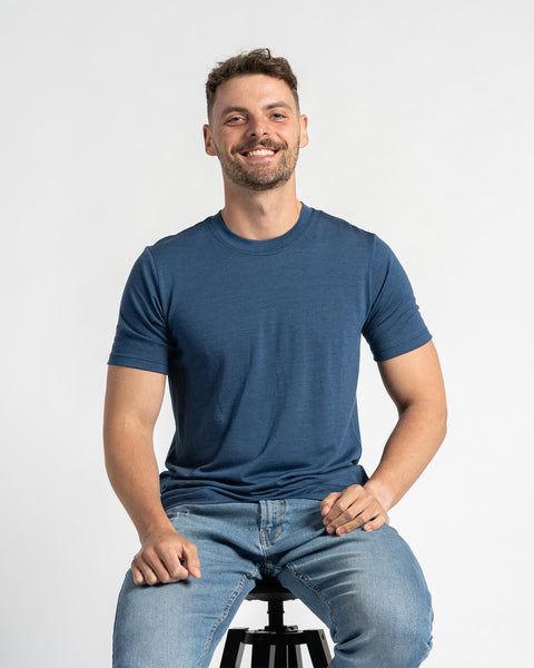 A man wearing a denim color t-shirt and light blue jeans sits on a stool against a light background. He has short brown hair, a light beard, and is smiling. The menique merino wool t-shirt is the primary focus, showcasing its fit and appearance while the man is seated.