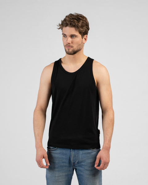A studio portrait of a man looking off to the left side of the frame with his arms relaxed by his sides. He is wearing a black tank top and blue jeans against a light grey background.