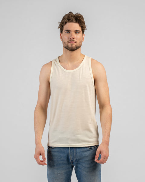 A studio portrait of a man standing and looking directly at the camera with his arms relaxed by his sides. He has wavy brown hair and a short beard. He is wearing a light beige, scoop-neck tank top and medium-wash blue jeans against a plain light grey background.