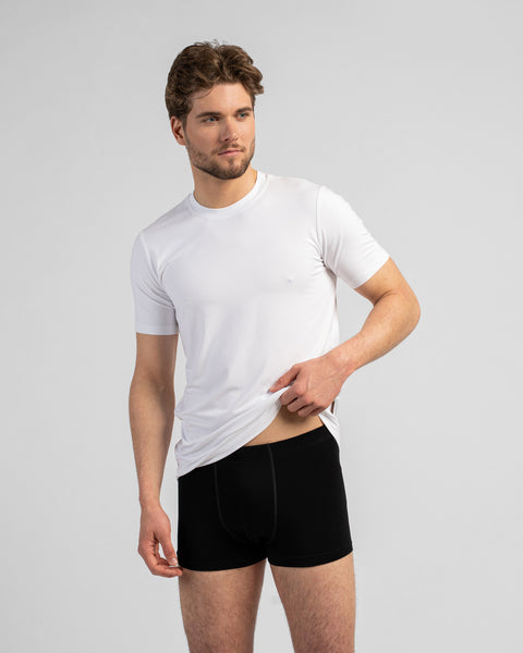 A studio portrait of a man looking to the side while wearing a white t-shirt and black boxer briefs. He is using one hand to gently lift the hem of his shirt against a plain light grey background.