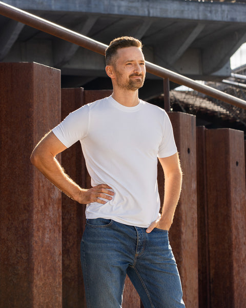 A man stands outdoors against a backdrop of rusted steel beams and a modern concrete overpass. He is wearing a plain white short-sleeved t-shirt and blue jeans. His right hand rests on his hip, and his left hand is tucked into his front pocket as he looks off into the distance.