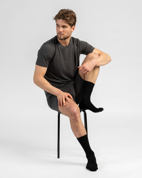 A studio portrait of a man sitting on a black stool, looking off to his right against a plain light grey background. He is wearing a matching set of charcoal grey short-sleeved t-shirt and long boxer briefs, paired with black crew socks.