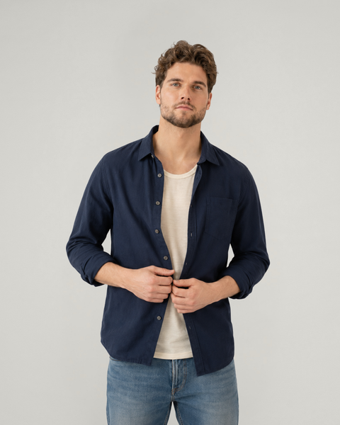 A man with wavy brown hair and a short beard stands against a plain light grey background, looking directly at the camera. He is wearing a light natural-beige scoop-neck top underneath an unbuttoned dark navy blue long-sleeved shirt, paired with medium-wash blue jeans. His hands are resting gently on the lower edges of the open shirt.
