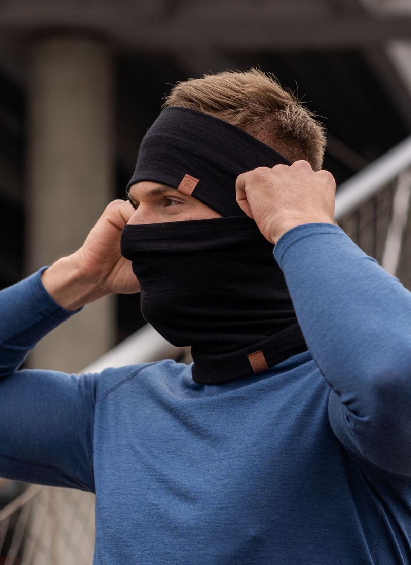 A close-up shot of a man outdoors with a blurred background of stairs. He is wearing a blue long-sleeved top, a black headband, and a black neck gaiter pulled up to cover his mouth and nose. He has his hands raised, adjusting the sides of the gaiter. Both the headband and the gaiter feature a small, square brown logo patch.