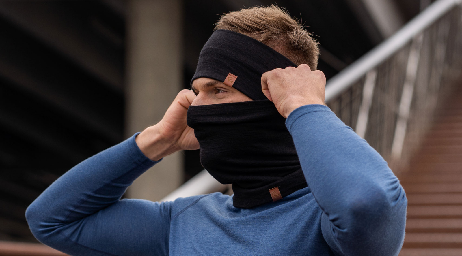 A close-up shot of a man outdoors with a blurred background of stairs. He is wearing a blue long-sleeved top, a black headband, and a black neck gaiter pulled up to cover his mouth and nose. He has his hands raised, adjusting the sides of the gaiter. Both the headband and the gaiter feature a small, square brown logo patch.