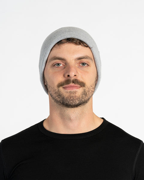 A person with light skin, short brown hair, a mustache, and light-colored eyes is shown in a headshot against a white background. They are wearing a light gray knit beanie with a small brown tag on the side and a black long-sleeved shirt. They are looking directly at the camera.