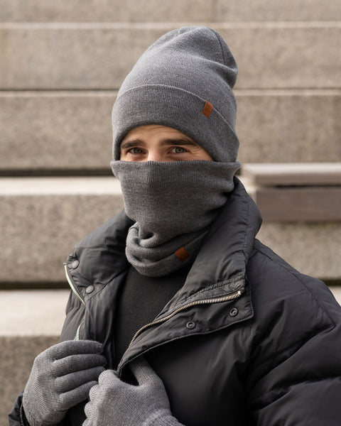 A person, who appears to be male, is standing outdoors on concrete steps, wearing a dark gray knit beanie, a dark gray knit neck gaiter pulled up over their mouth and nose, dark gray knit gloves, and a black puffer jacket. Only their eyes are visible above the neck gaiter. The beanie has a small brown tag on the folded brim. The person seems to be looking directly at the camera.