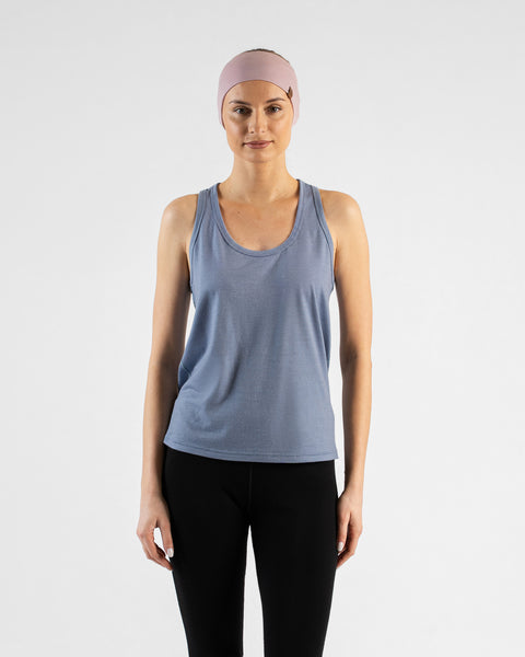 A studio portrait of a blonde woman standing and looking directly at the camera against a plain light grey background. She is wearing a dusty blue sleeveless top, black leggings, and a wide dusty pink headband featuring a small brown logo tag.