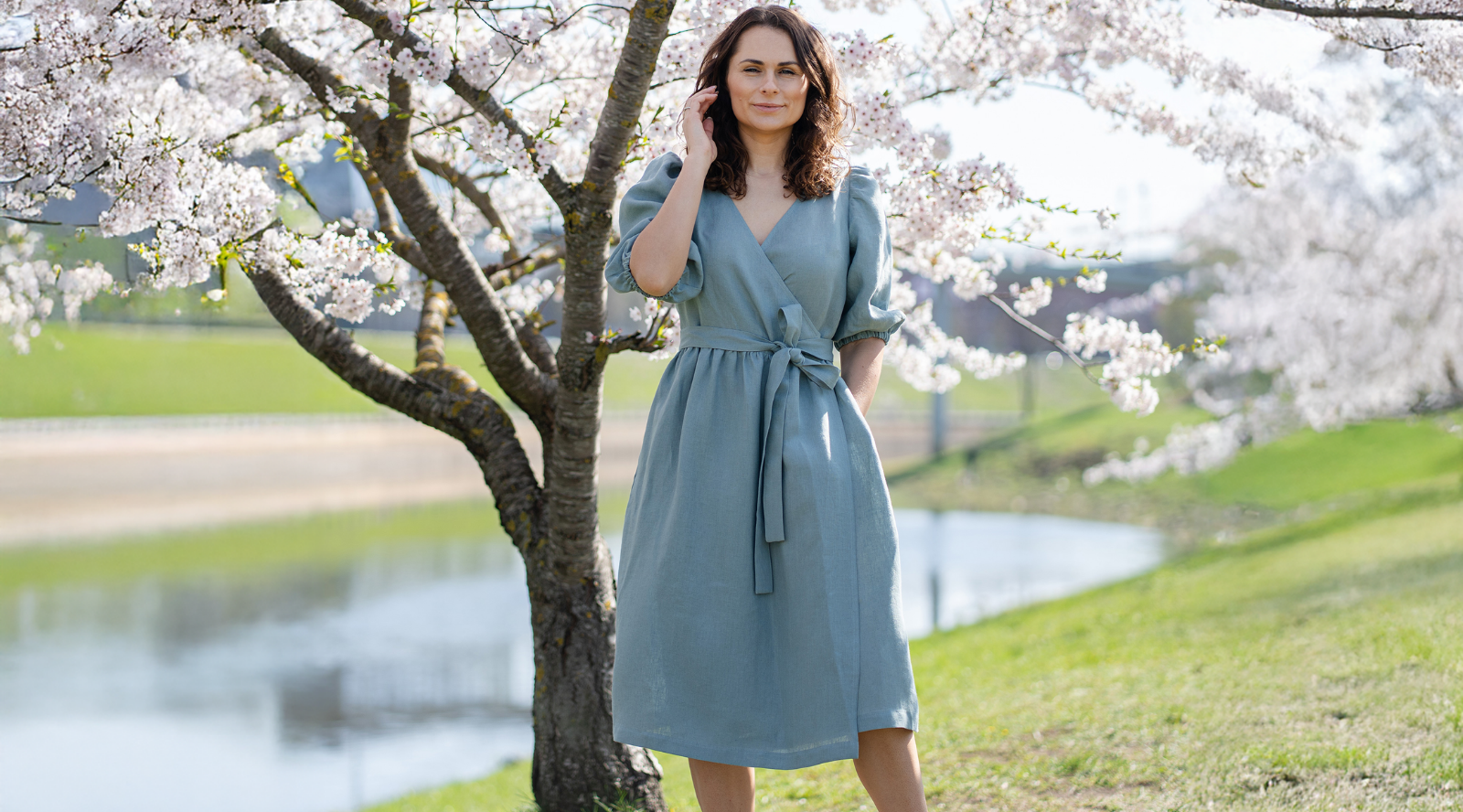 Woman wearing a blue linen midi wrap dress standing outdoors by a pond under blooming cherry blossom trees on a sunny spring day.